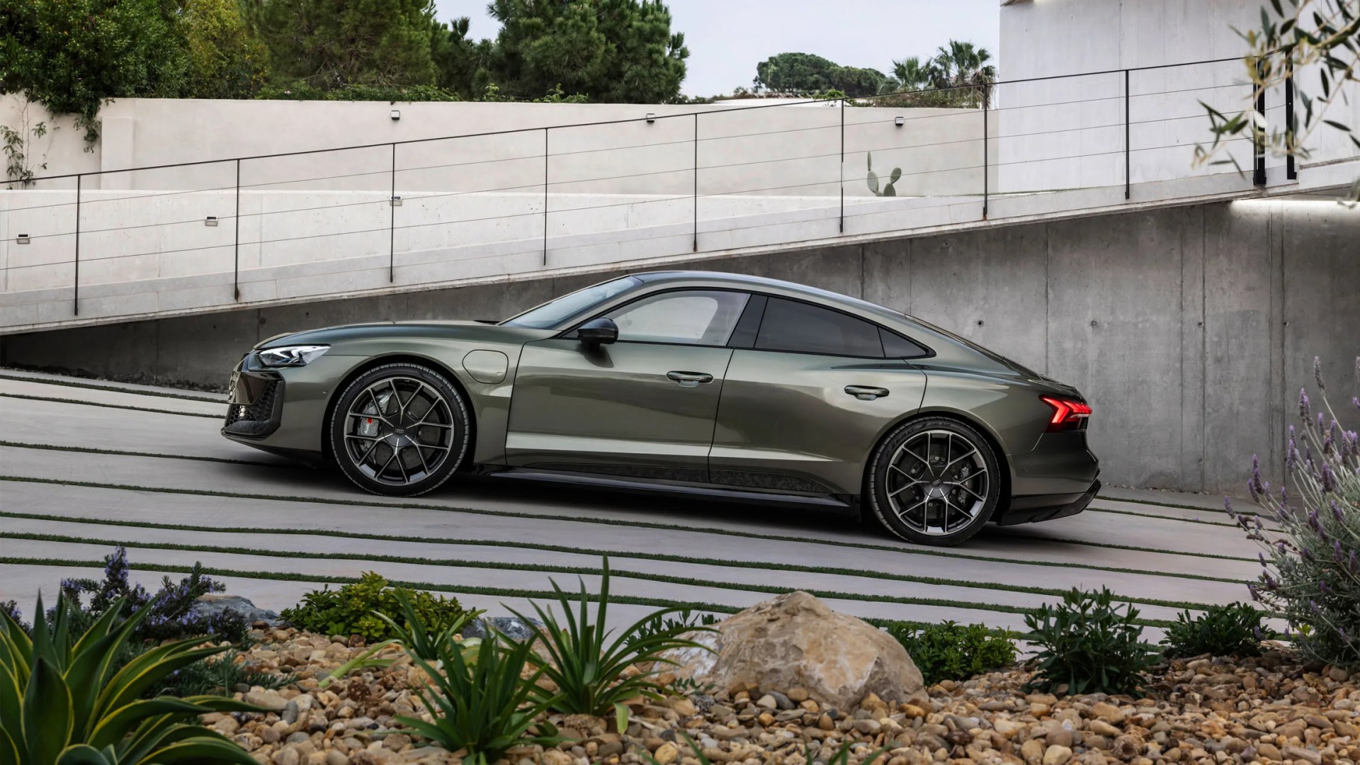 Audi RS e-tron GT Performance Edition driving up the hill towards the left edge of the image. Plants and rocks are the foreground. In the background is a modern looking ramp to a building with a metal and glass side rail.