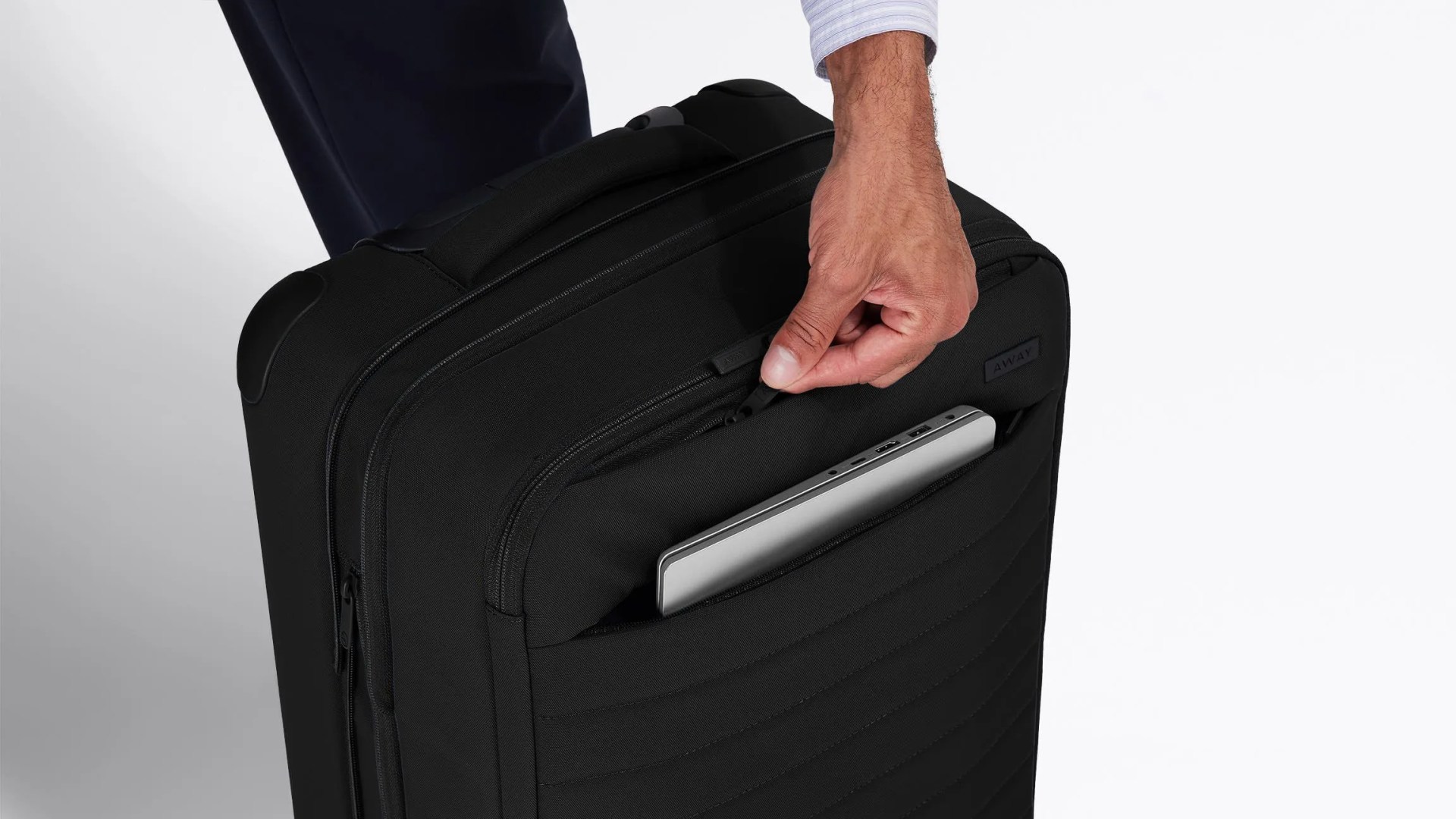 Close up shot of a hand reaching zipping the external zipper pocket of a black piece of Away soft-sided luggage to reveal a silver laptop inside the pocket