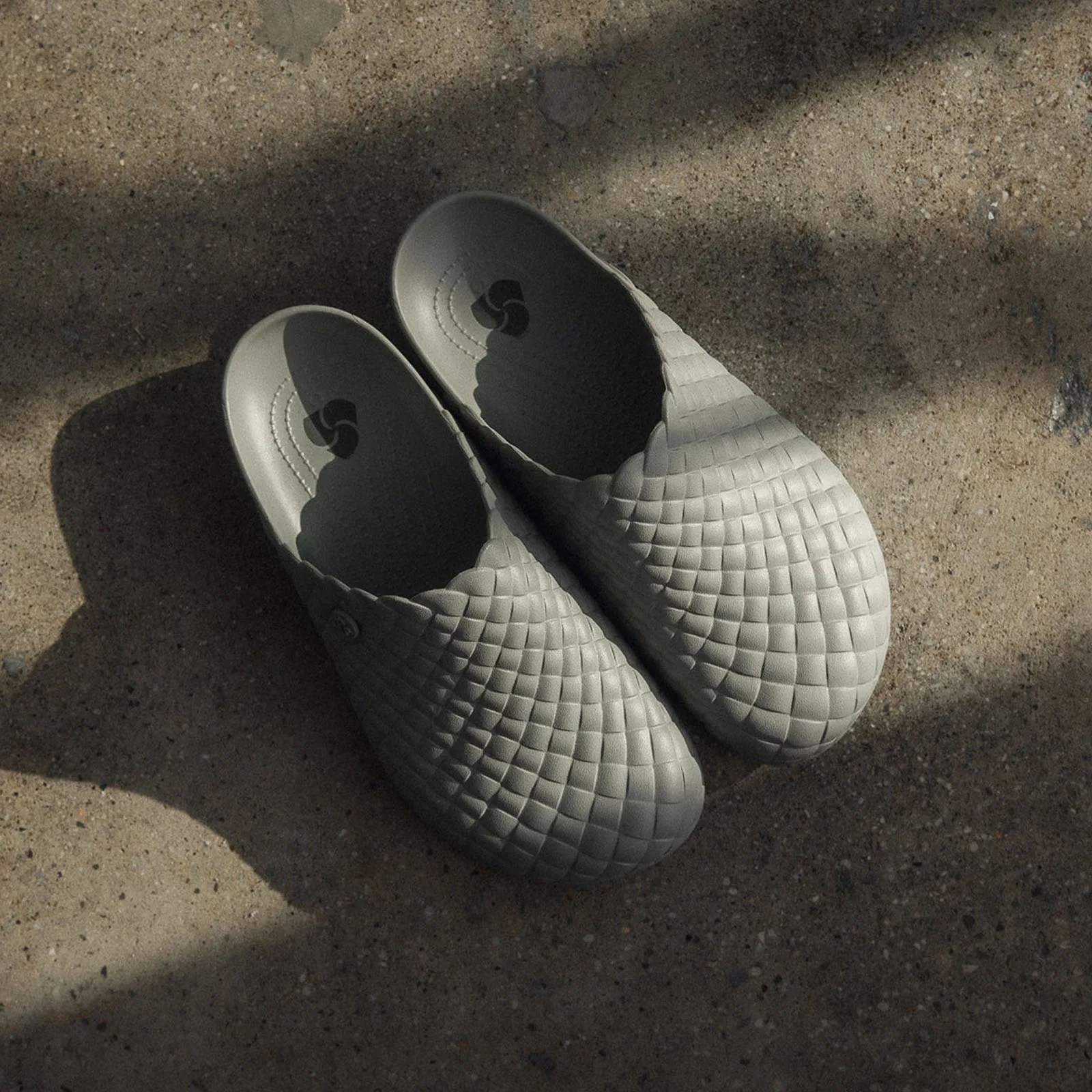 A pair of Crocs x 3sixteen woven Dylan clogs positioned diagonally with the toes pointing to the lower right corner of the image. They are resting on a concrete colored floor with various shadows casting over them.