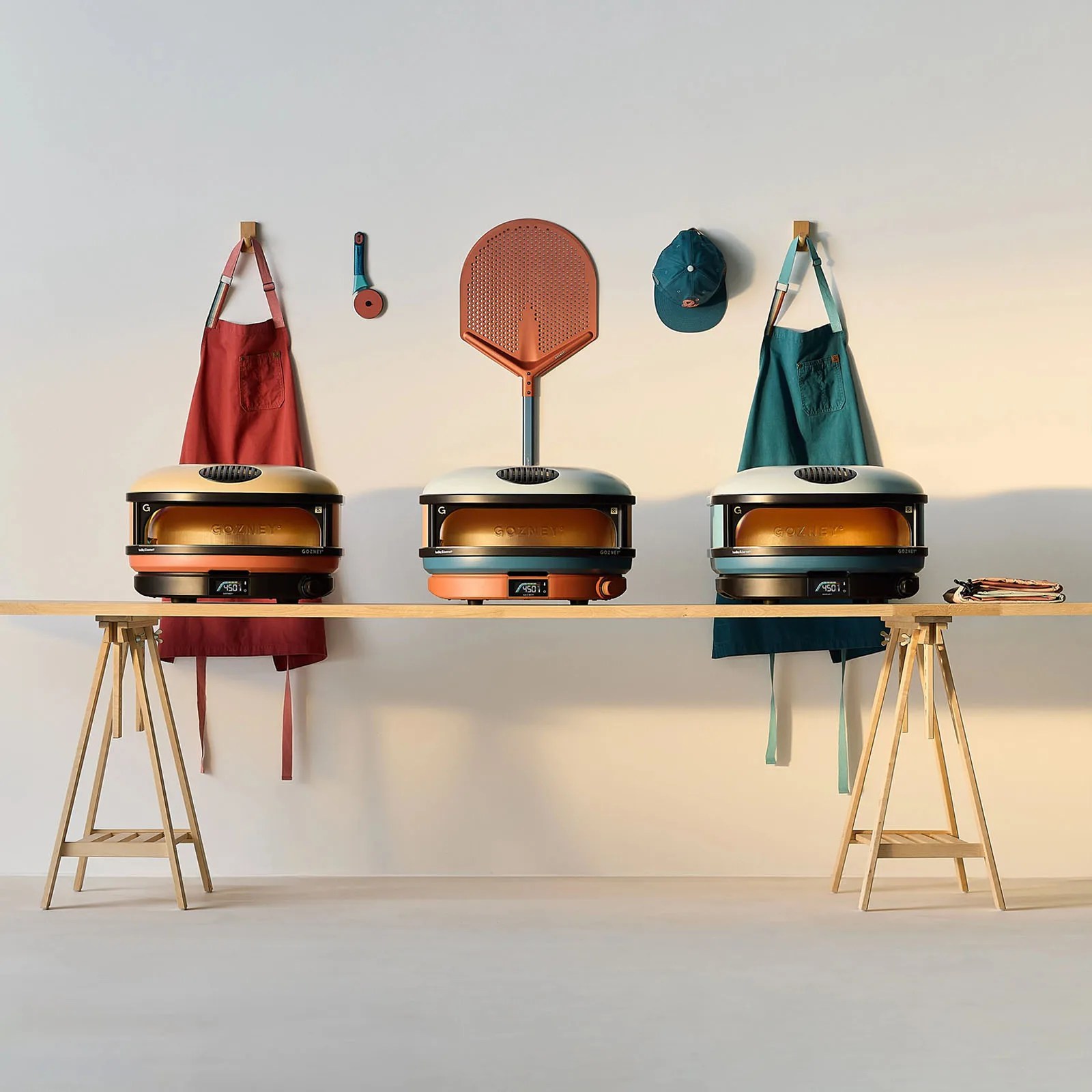 Three Gozney x Hedley and Bennett limited edition pizza ovens sitting on a wooden table with custom aprons in background