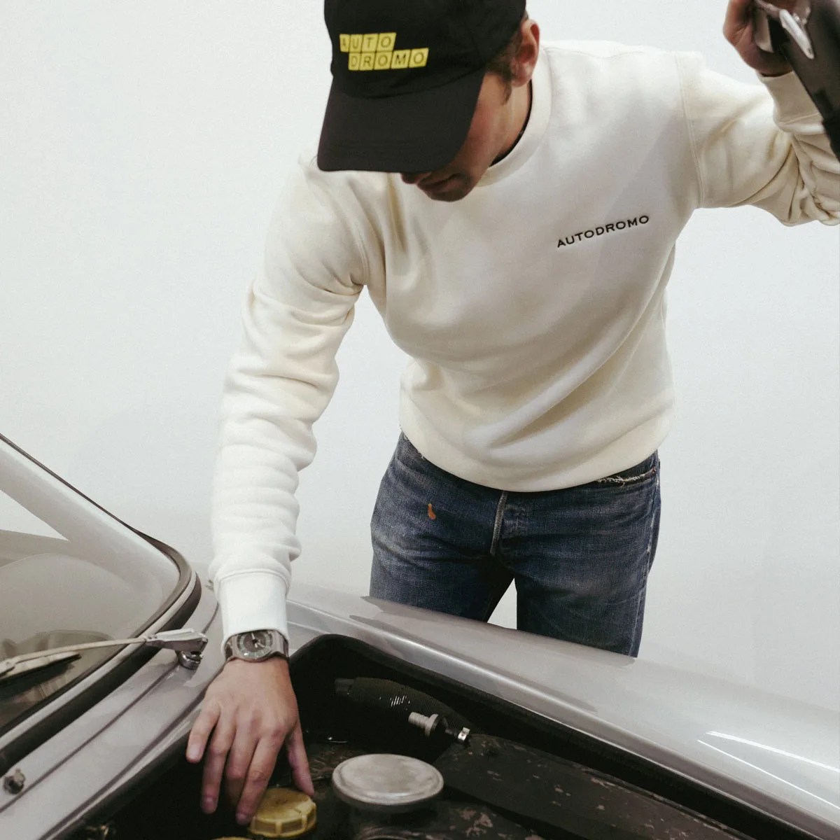 A young man wearing off white colored sweatshirt with black Autodromo brand on the front breast looking at the engine bay of a vintage silver car