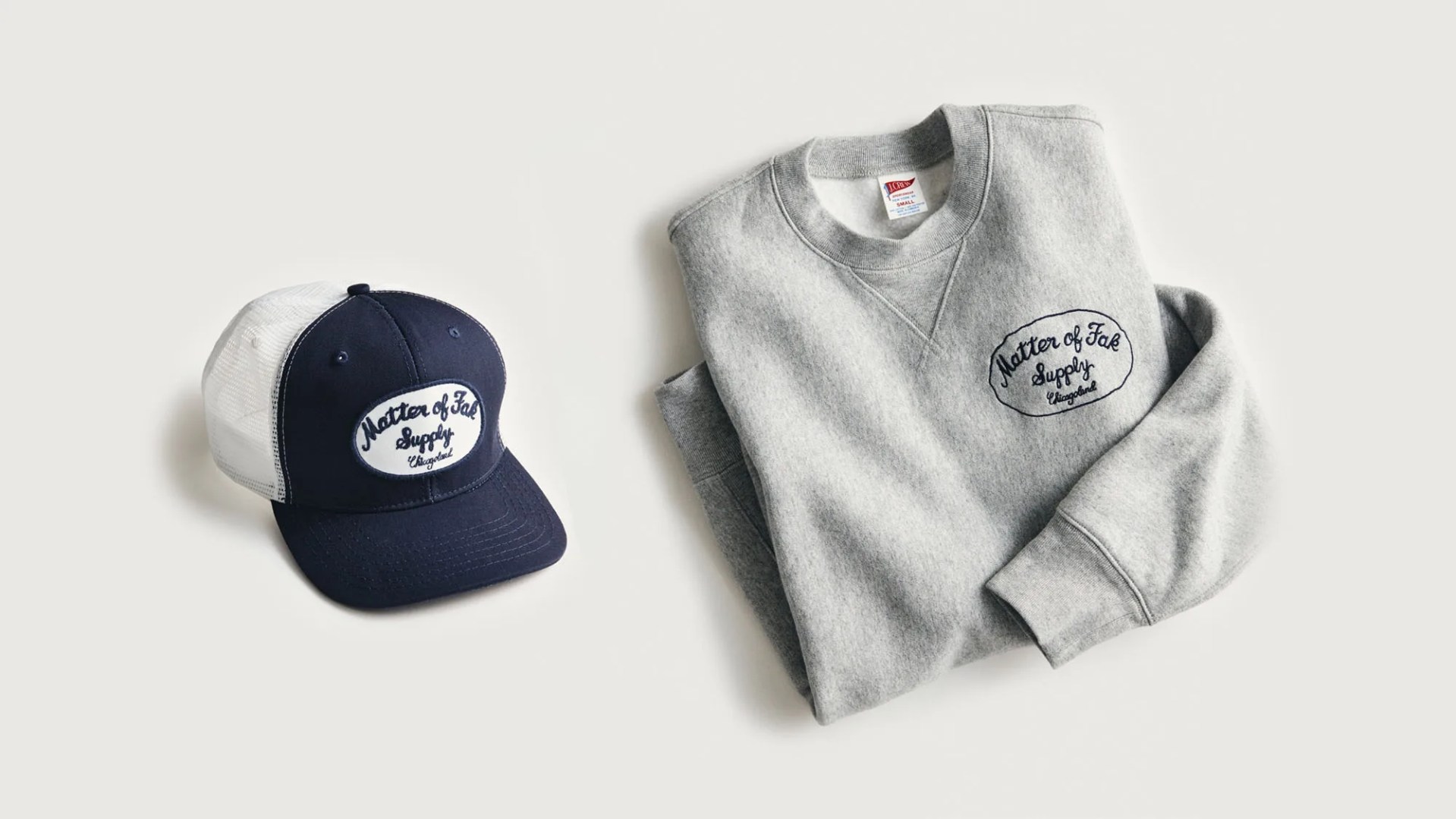 A blue and white trucker baseball hat sitting to the left of a gray folded sweatshirt made by J. Crew that each gesture a Matter of Fak Supply Logo from the fit FX TV Show The Bear