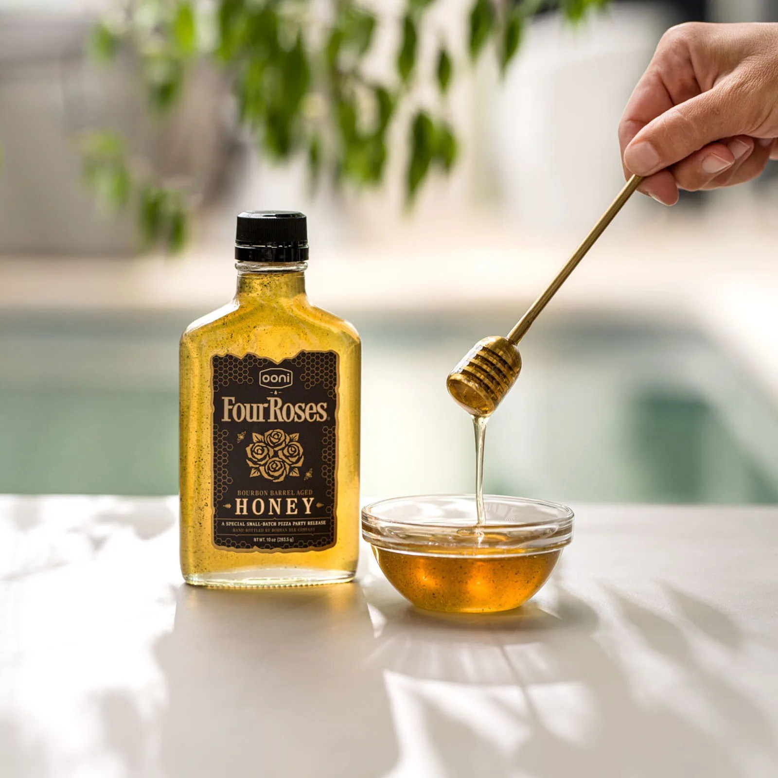 A bottle of Ooni x Four Roses honey on an outdoor table with a pool in the background. Next to the bottle, a hand is holding a wooden spoon that’s been dipped in the honey with a long drip hanging off the spoon falling into a glass bowl filled with honey.