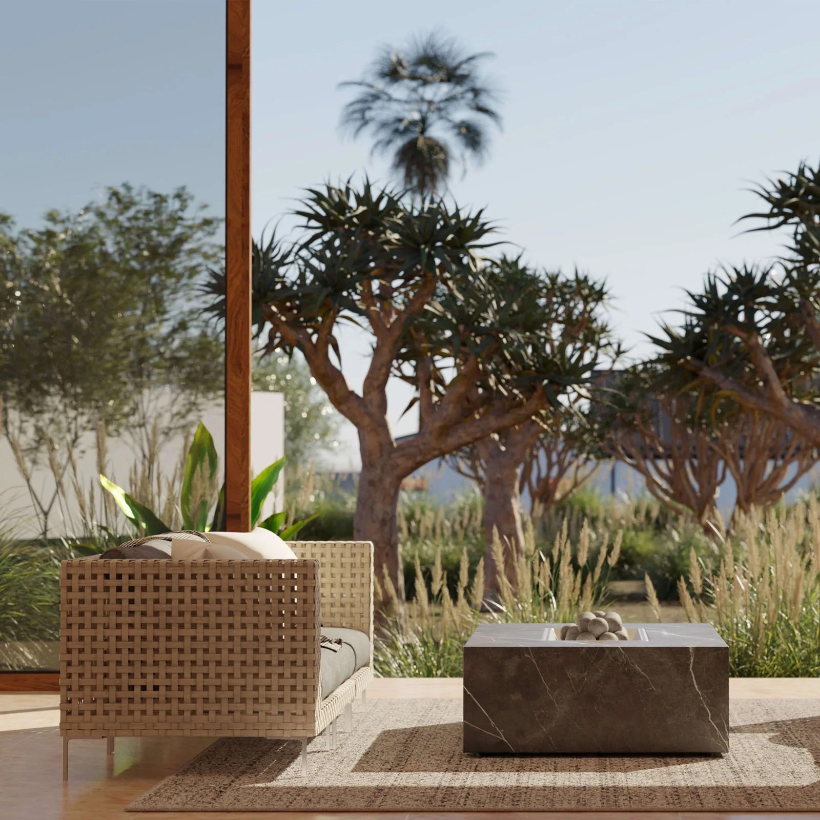 Outer open weave wicker love seat viewed from side next to a square stone fire pit with a variety of trees, tall grasses and blue sky in the backdrop.