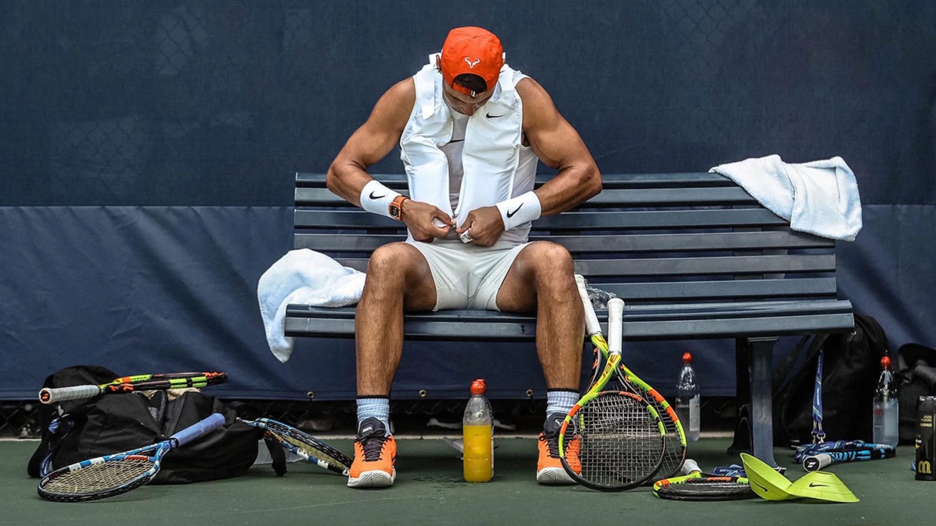 Pro Tennis star Rapha Nadal putting on a white cooling vest designed by Nike packed with ice during the 2018 US Open while sitting on a sideline bench surrounded by tennis gear.