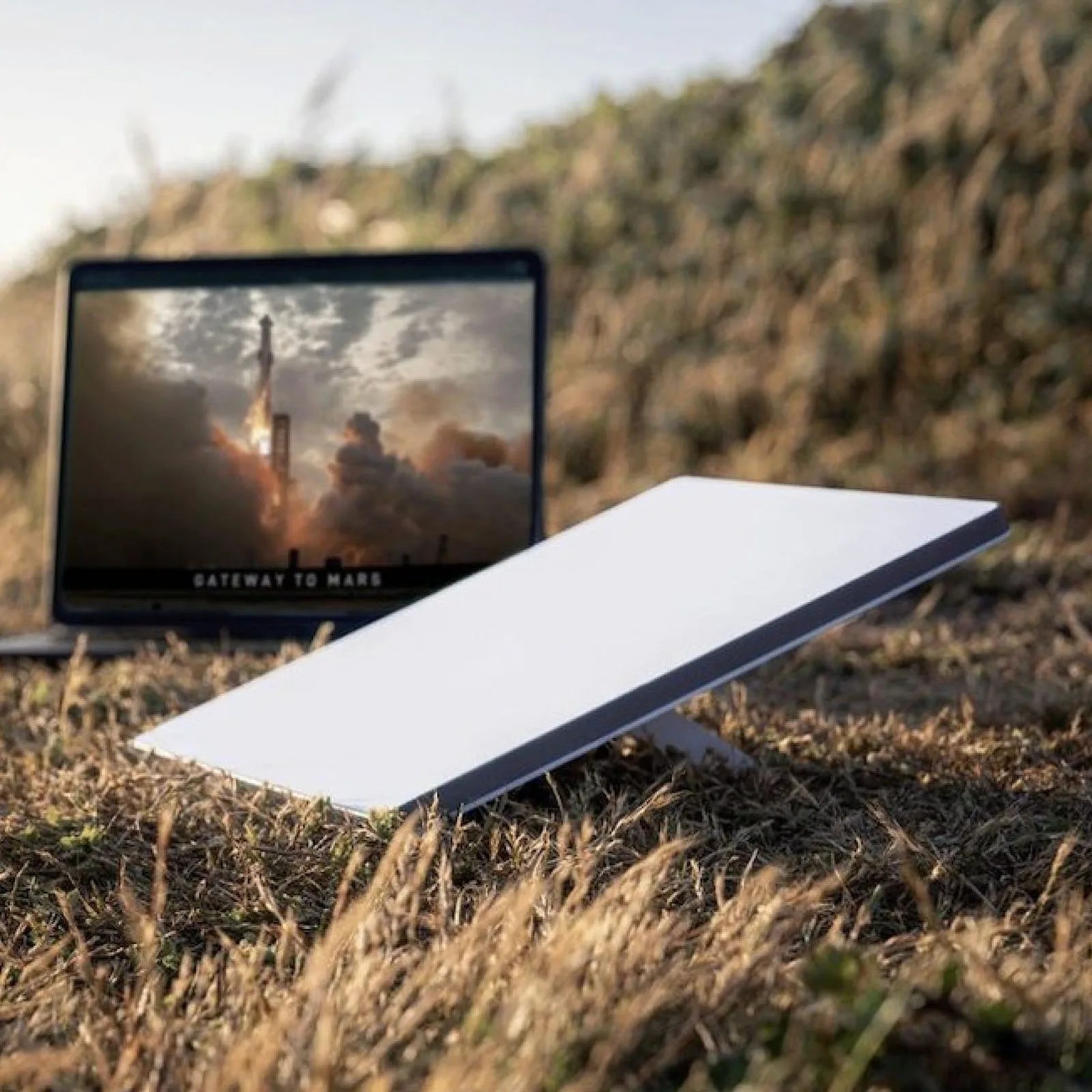 A Starlink mini resting at an angle on its kickstand in a grassy field with a blurred laptop screen in the background.