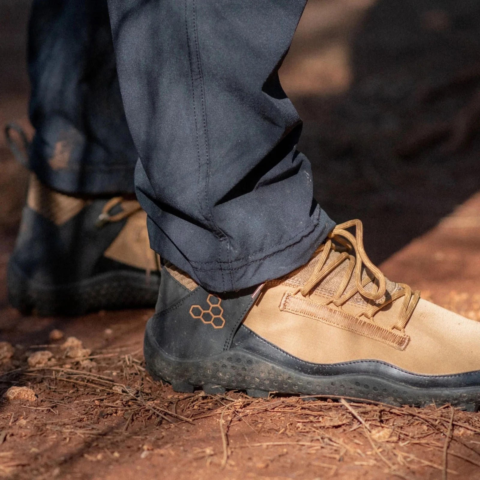 A close up ankle shot of Vivobarefoot Magna Lite boots on a person wearing navy pants photographed from side on a dirt trail