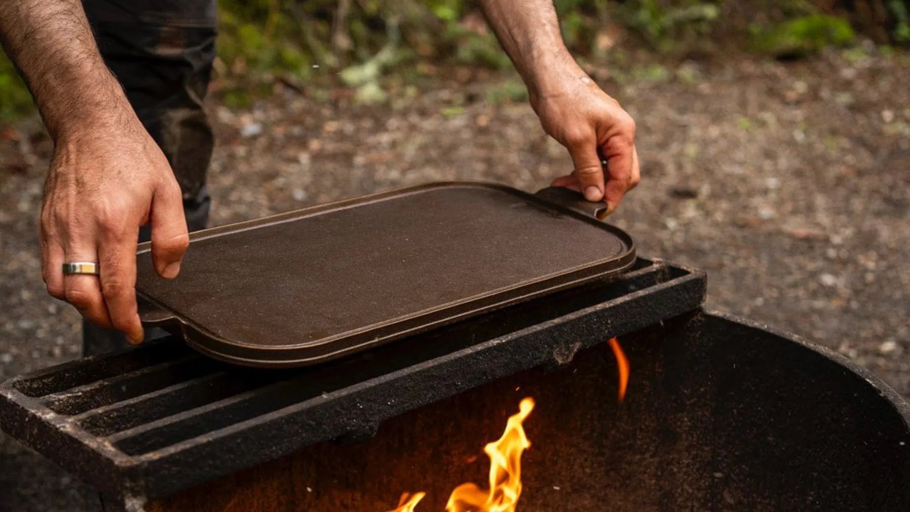 man holding a cast-iron griddle over a fire