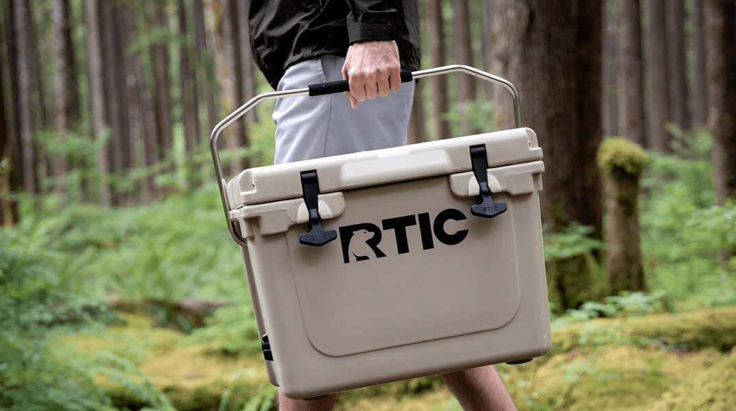 A tan colored RTIC 20 quart cooler being held in hand by someone walking across a forest